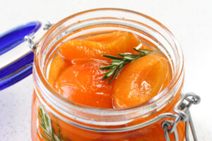 An open jar of poached apricots in syrup with a sprig of rosemary