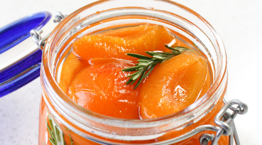 An open jar of poached apricots in syrup with a sprig of rosemary