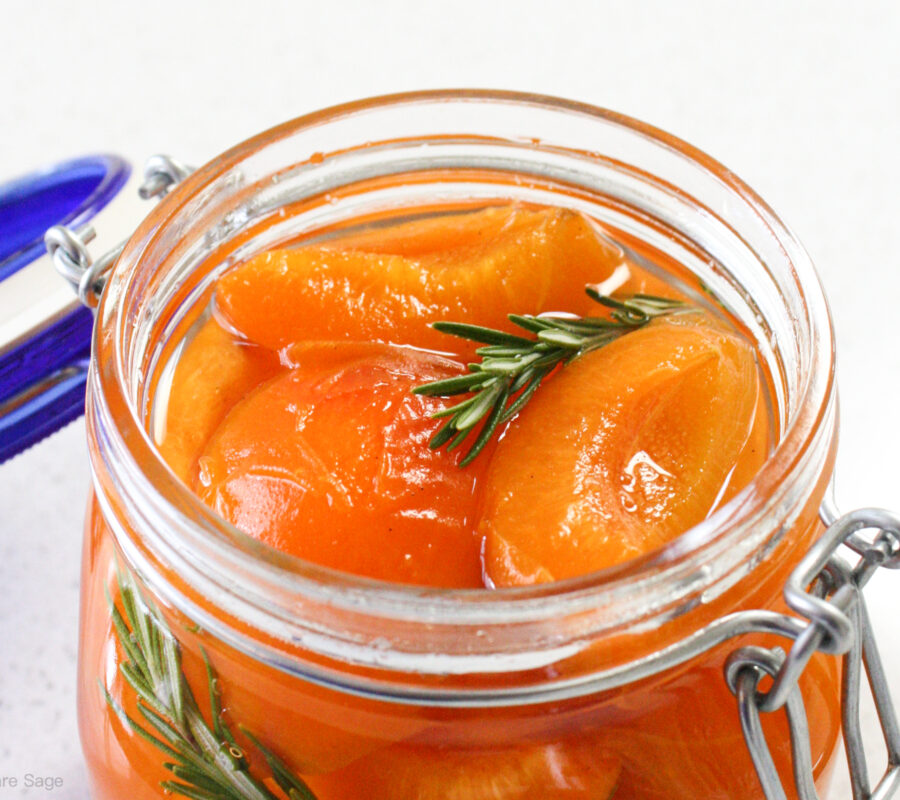 An open jar of poached apricots in syrup with a sprig of rosemary