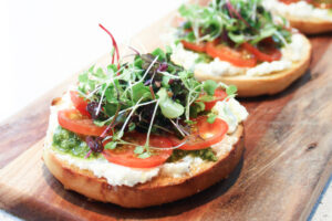 A close up view of a toasted bagel topped with ricotta, pesto, tomato and micro herbs