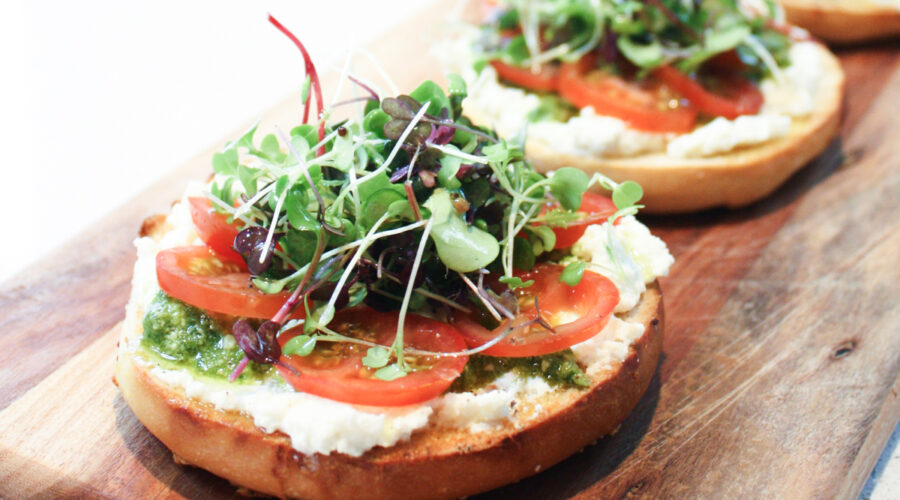A close up view of a toasted bagel topped with ricotta, pesto, tomato and micro herbs