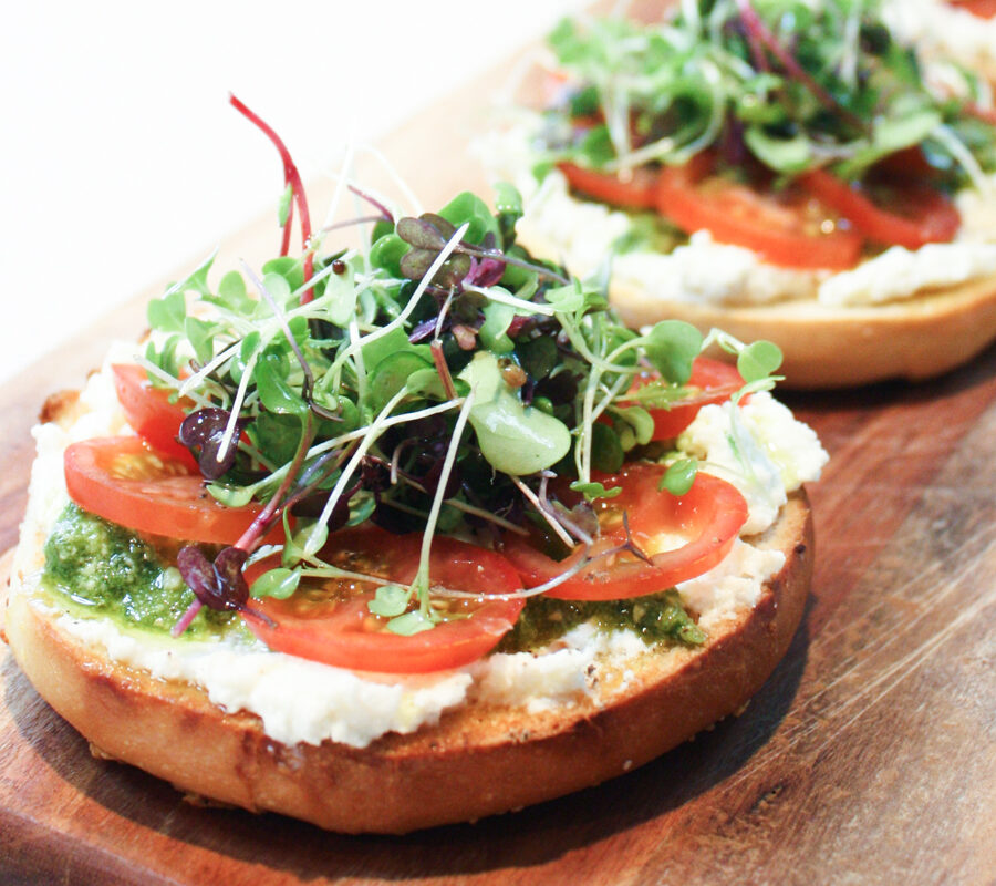 A close up view of a toasted bagel topped with ricotta, pesto, tomato and micro herbs